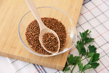small grains of brown natural dry buckwheat in a transparent glass plate and in a wooden spoon
