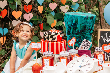 Little smiling girl 2 years old dressed as Alice in Wonderland near table with fairy tale props. Cake, Hatters hat, inscriptions Eat me and Drink me, clock, card suits. Themed childrens book birthday