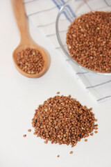 small grains of brown natural dry buckwheat in a transparent glass plate and in a wooden spoon