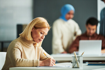 Serious mature blond teacher in beige pullover checking grammar test while sitting by desk in front of camera against two students