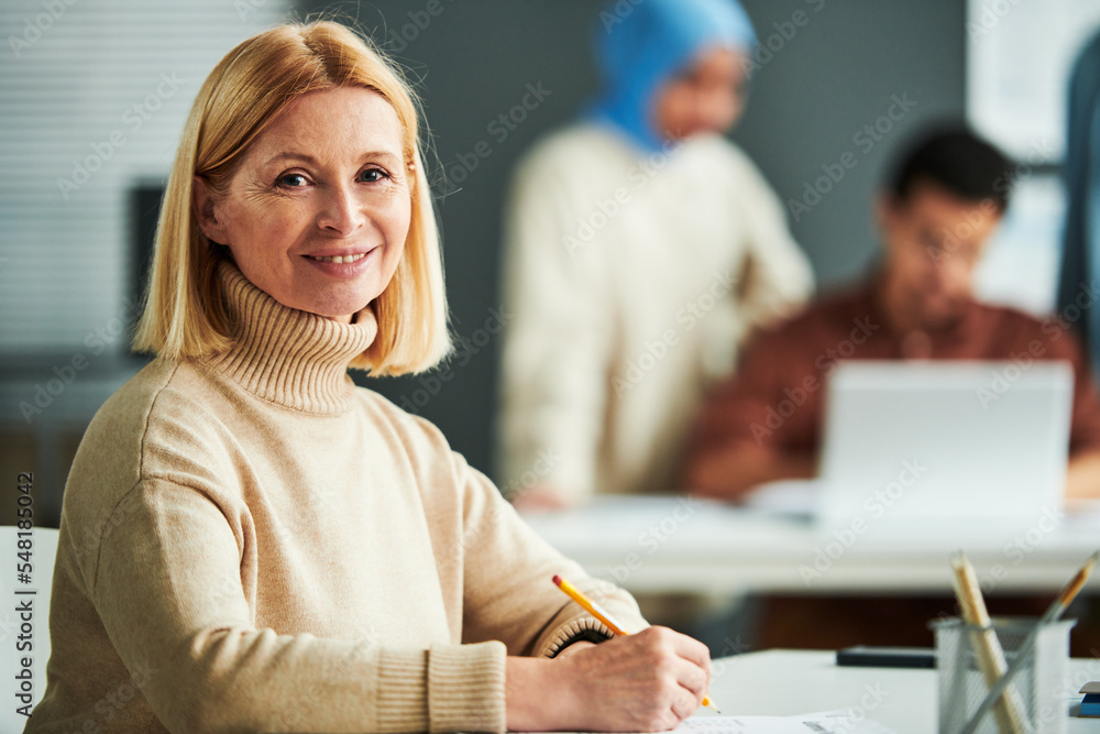 Aufkleber successful experienced teacher with blond hair looking at camera while sitting by desk in classroom 