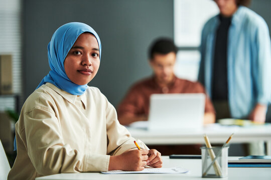 Young Muslim woman in blue hijab looking at camera while lchecking or carrying out grammar test by workplace against two guys