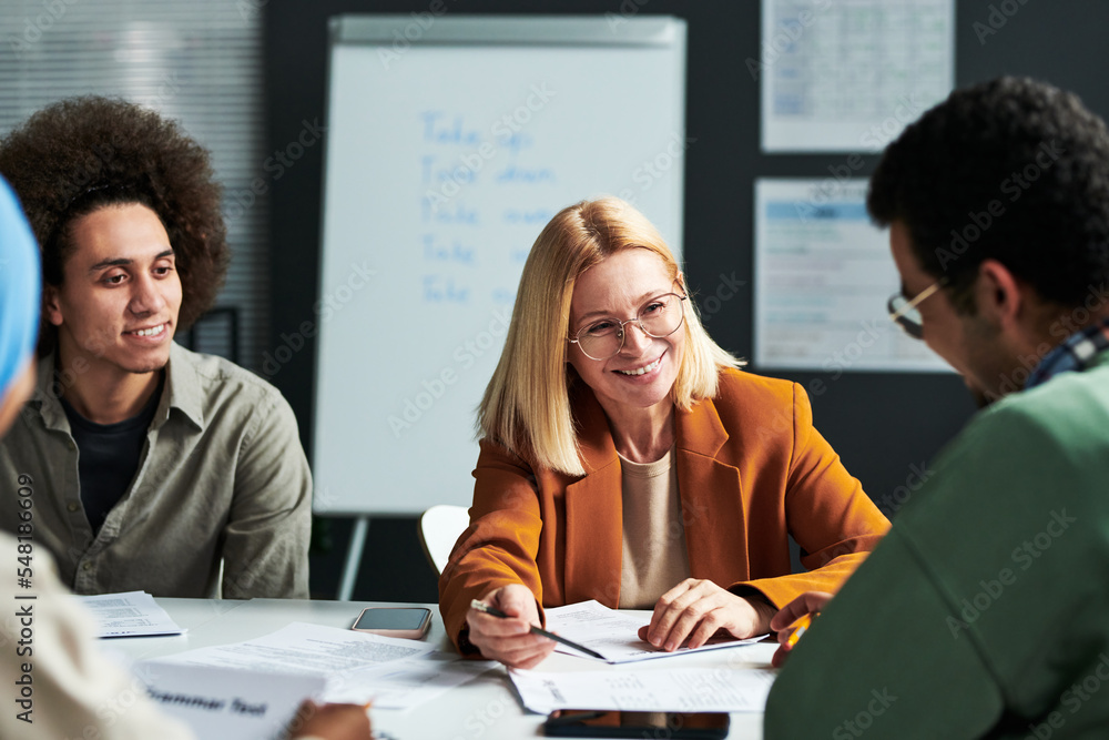 Poster happy mature blond teacher of linguistics explaining something to one of students while looking at h