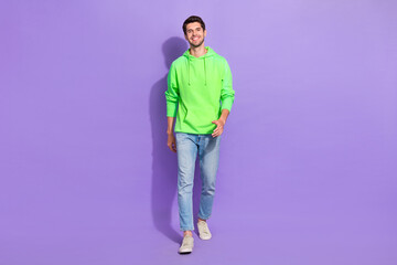 Fototapeta na wymiar Full size photo of handsome cheerful positive guy dressed green hoodie denim jeans walking street isolated on violet color background