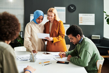 Mature blond teacher showing document with English grammar test to Muslim female student in hijab while both standing by workplace - obrazy, fototapety, plakaty