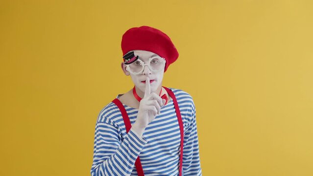 The guy mime in a striped and bright costume and makeup brings his index finger to his lips and shows a shh sign looking at the camera. Pantomime makes a performance on a yellow background