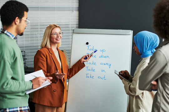 Confident mature teacher of English language pointing at whiteboard with list of phrasal verbs and explaining their meaning to students