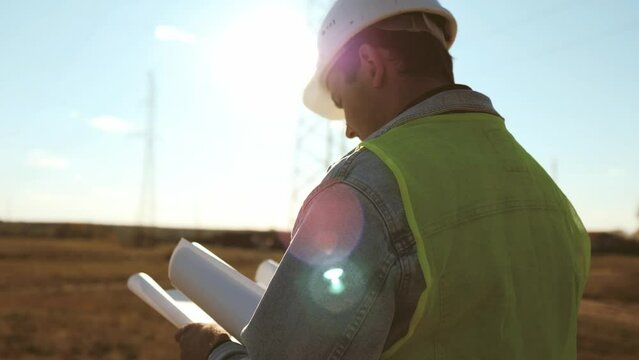 Worker male engineer using documents for checking data while standing against high voltage power towers. Power engineering specialist with working documentation working near electric poles.