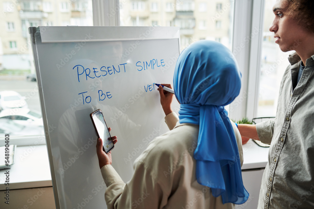 Poster back view of young muslim female teacher explaining multi-ethnic guy present simple tense while wiri