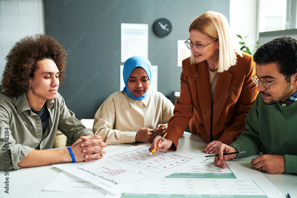 Poster Confident blond teacher in eyeglasses looking at one of students while explaining information in document at lesson of foreign language