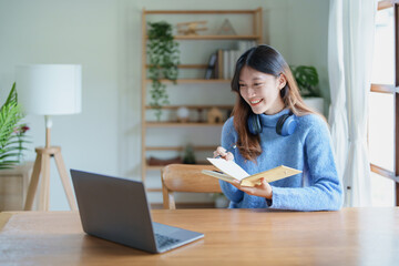 Portrait of a teenage Asian woman using a computer, wearing headphones and using a notebook to study online via video conferencing on a wooden desk in library