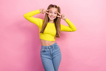 Portrait of optimistic girl with straight hair dressed yellow long sleeve showing v-sign near eyes isolated on yellow color background