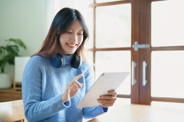 Portrait of a teenage Asian woman using a tablet, wearing headphones to study online via video conferencing on a wooden desk in library