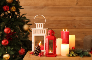 Christmas lanterns with burning candles, fir branches and cones on table near wooden wall