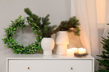 Vases with fir branches, Christmas mistletoe wreath and candles on chest of drawers in bedroom