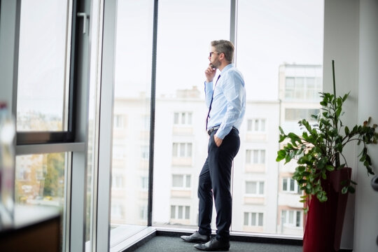 Full Length Of Thinking Businessman Looking Out Of Office Over City. Confident Man Wearing Glasses And Business Casual.