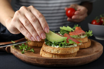 Woman adding avocado onto tasty rusks with salmon and cream cheese at black table, closeup