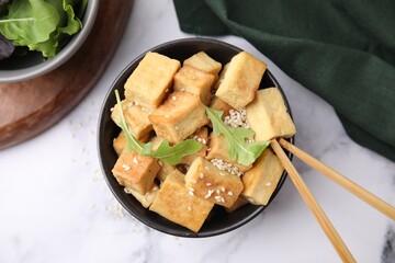 Delicious fried tofu with basil and sesame seeds in bowl on white marble table, flat lay
