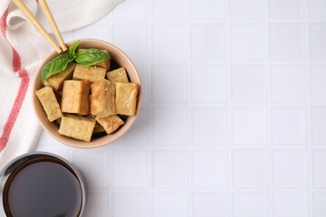 Delicious fried tofu with basil and sesame seeds in bowl on white tiled table, flat lay. Space for text