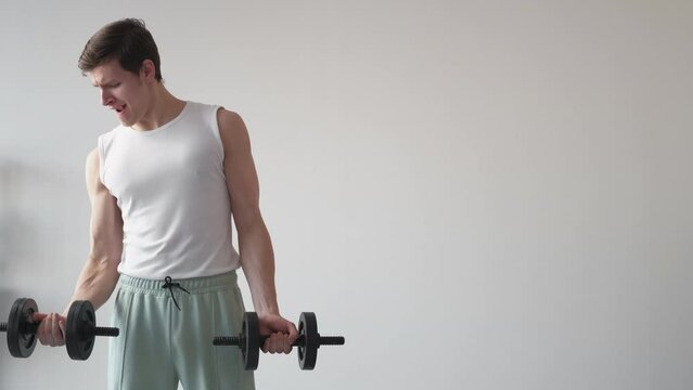 Hard sport. Powerless man. Home training. Tense slender guy feeling difficulties doing workout with dumbbells in light room interior copy space.