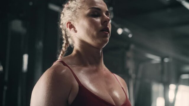 A Resilient Young Sports Woman Training Using Weights and Barbell in a Dark Gym. A Female Athlete Smiles While Doing her Daily Strength Workout and Exercises. Handheld Slow Motion Shot
