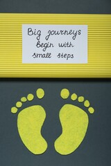 Card with phrase Big Journeys Begin With Small Steps and paper feet on dark background, top view....