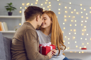 Happy couple in love sitting on sofa at home, holding gift box, smiling and touching noses. Young woman kissing boyfriend and thanking him for Saint Valentine's Day or relationship anniversary present