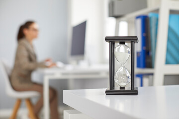 Black sandglass with pouring white sand at desk on office blur background with woman working on...