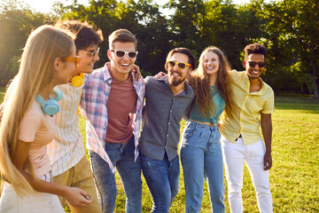 Young happy multiracial people men and woman walk around summer park putting their hands on each other's shoulders and enjoying spending time together with friends in sunny nature