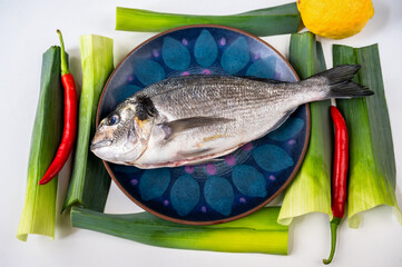 Part of raw sea bream (dorada, king bream) fish on plate, leaf, chilli, lemon on white.