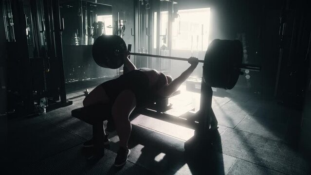 A Professional Bodybuilder Training by Lifting a Heavy Barbell and Pushing her Limits In a Dark Gym. Female Energetic Athlete Doing Core Exercise with Bench Press Workout. Slow Motion Zoom in Shot