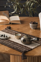 Fototapeta na wymiar Minimalist composition of living room interior with wooden round table, book, incense, bowl with nuts, patterned table pad, beige vase with leaves and personal accessories. Home decor. Template.