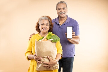 Happy senior indian couple hold grocery paper bags with food and blank smart phone screen mockup isolated on beige background. studio shot.