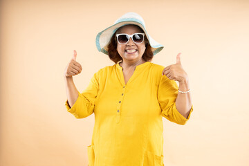 Happy mature indian woman wearing hat and sunglasses in summer do thumbs with both hands standing...