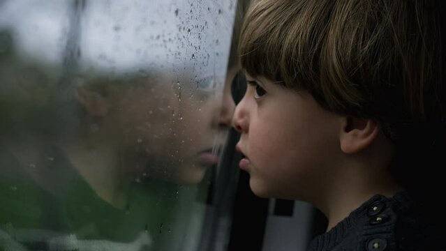 Melancholic little boy leaning on train window watching landscape passing during rainy day. Small child traveling
