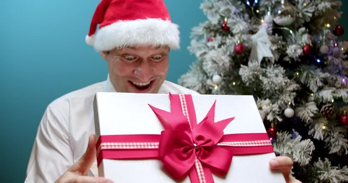 Happy smiling dad in Christmas hat Santa Claus receives a gift, rubs his hands before opening box with a gift, looks into box with surprise, happy face man delighted with surprise opens box with gifts