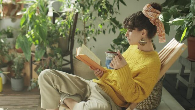 Medium shot of young woman drinking tea and reading book while resting in living room decorated with houseplants