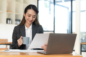 Beautiful young Asian businesswoman smiling holding a coffee mug and laptop working at the office.