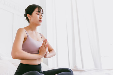 Fototapeta na wymiar Young attractive sporty Asian woman practicing yoga on the bed, doing Ardha Padmasana exercise, meditating in Half Lotus pose with namaste, indoor working out at home, wearing sportswear.
