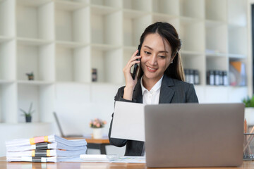 Fototapeta premium Smiling beautiful Asian businesswoman analyzing chart and graph showing changes on the market and holding smartphone at office.