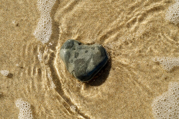Fototapeta na wymiar A heart-shaped stone on the shore. Sandy beach with a light sea wave. Background to a pleasant holiday, relaxing vacation and hiking in an ecologically clean area.