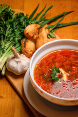 Bowl Of Traditional Borscht With Garlic And Herbs. A close-up of a red beet soup served with sour cream, garlic, herbs, and bread on a wooden board.