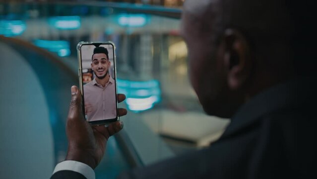 Back view unrecognizable African American businessman holding phone with talking Indian ethnic man worker entrepreneur on mobile screen multinational collaboration business chat video call conference