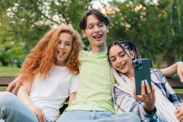 Fototapeta premium Portrait of a happy three teenagers sitting on the bench and making photo on smartphone. Relaxing on the bench in university campus. Sunny summer day.