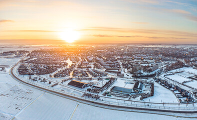 The yellow, golden, rising sun illuminates a small city in winter conditions. The houses, meadows and streets are covered in white snow, the water is frozen.