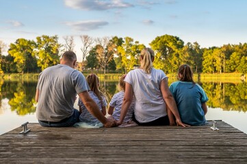 Lovely family with three kids sitting on a wooden dock over a lake on a sunny day - obrazy, fototapety, plakaty