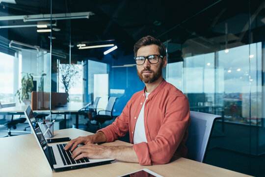 Portrait of a successful programmer inside the office, a man with a beard, glasses and a red shirt looks at the camera, a developer engineer uses a laptop to write code in a programming framework.