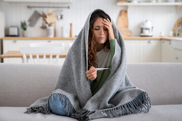 Sick exhausted woman with thermometer to measure body temperature sit on sofa at home wrapped in...