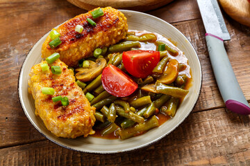 Cutlets with green beans and mushrooms in tomato sauce in a plate garnished with cherry tomatoes and green onions next to a knife.