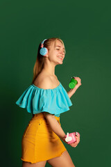 Music, dance. Portrait of young beautiful girl, student in summer outfit posing isolated over green background. Positive emotions, happiness, hobbies and joy